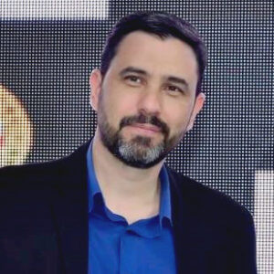 Daniel Escobar, homem branco, cabelo escuro e barba grisalha, veste paletó preto e camisa azul, olhando para a câmera